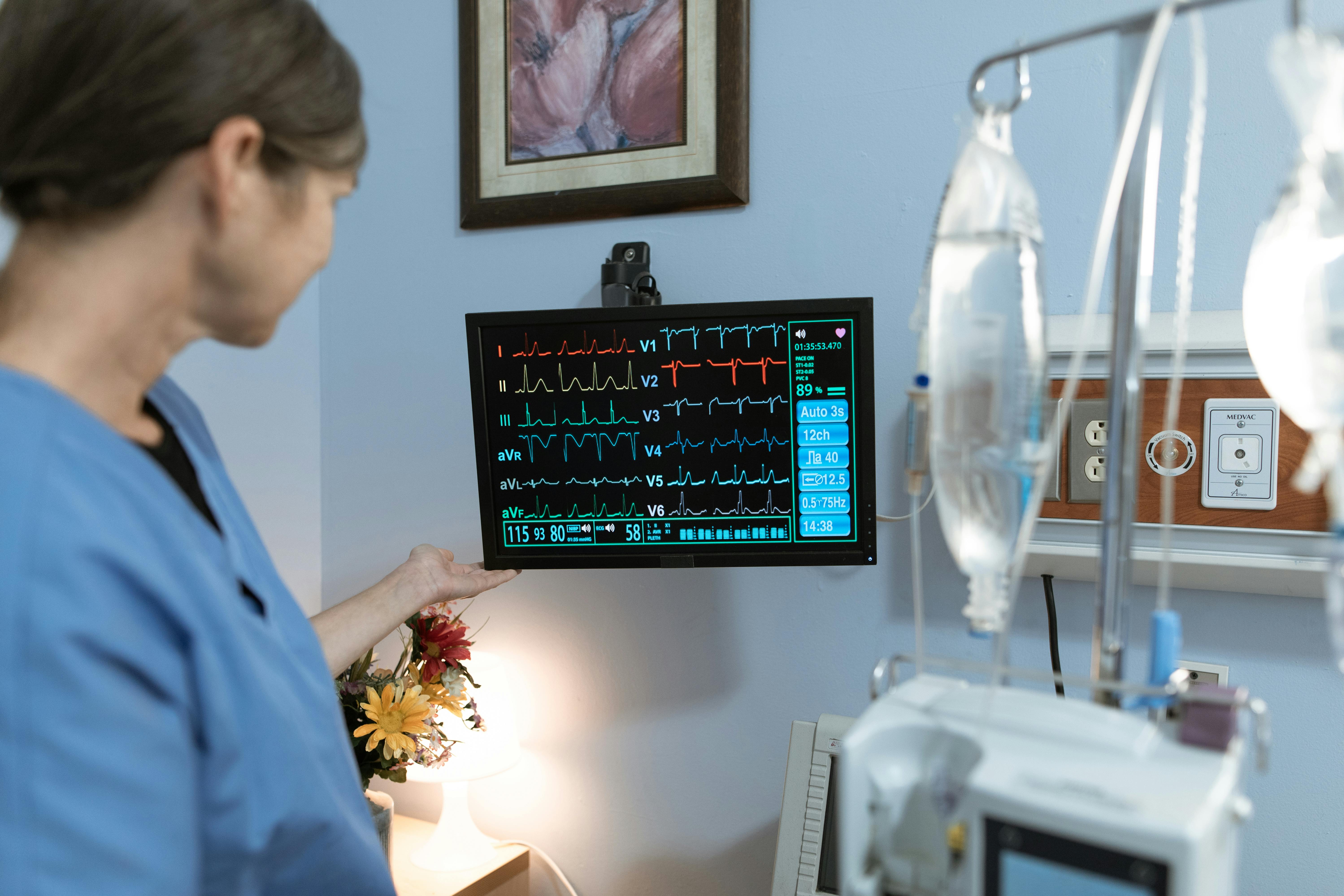 A nurse in blue scrubs examines a medical monitor displaying vital signs in a hospital setting.
